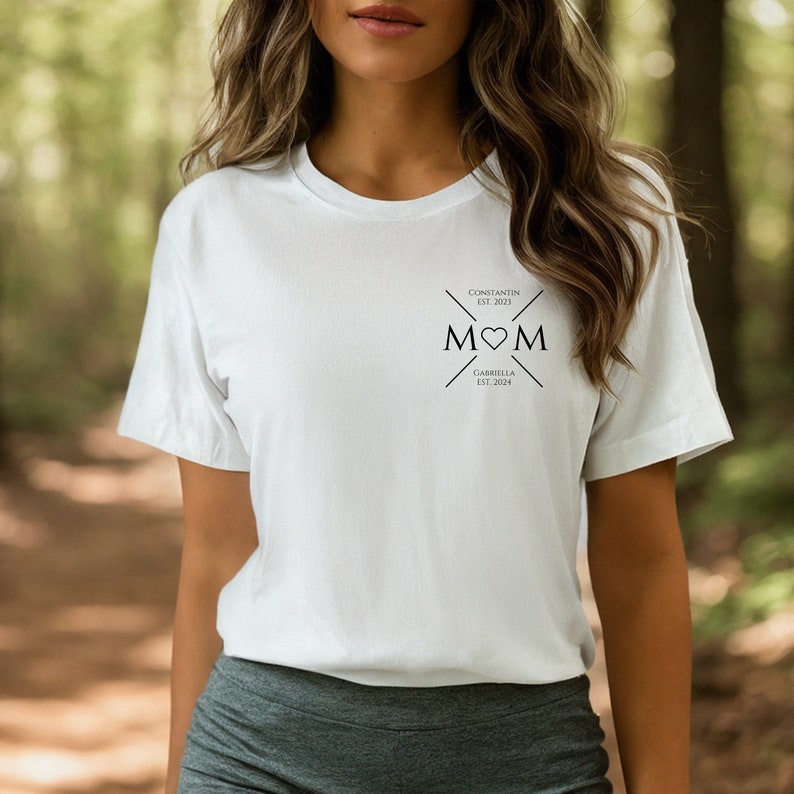 May include: White t-shirt with a black graphic design that says "MOM" with a heart in the middle. The graphic also includes the text "CONSTANTIN EST. 2023" and "GABRIELLA EST. 2024".