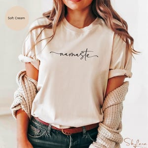 K&ouml;nnte beinhalten: Ein T-Shirt in zartem Cremeton mit dem Wort "namaste" in schwarzer Kursivschrift.
