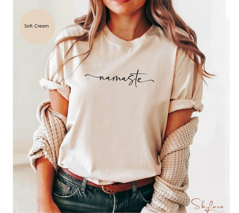 K&ouml;nnte beinhalten: Ein T-Shirt in zartem Cremeton mit dem Wort "namaste" in schwarzer Kursivschrift.