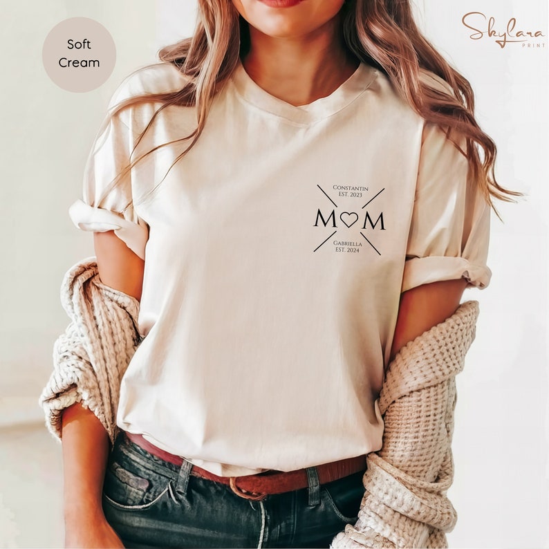 May include: A soft cream t-shirt with a black graphic design that says "MOM" with a heart in the middle. The graphic also includes the text "CONSTANTIN EST. 2023" and "GABRIELLA EST. 2024".