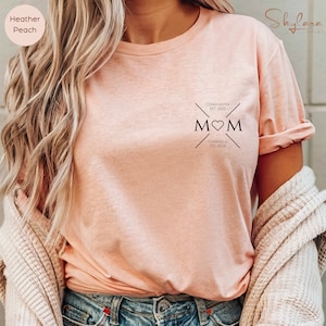 May include: A light pink t-shirt with a black graphic design that says "MOM" with a heart in the middle. The graphic also includes the text "Constantin Est. 2023" and "Gabriella Est. 2024".
