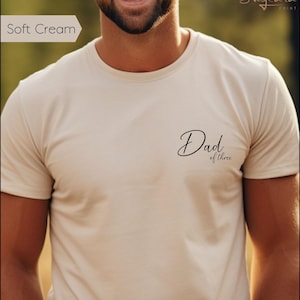 Può includere: T-shirt color crema chiaro con la scritta "Dad of three" in corsivo nero. La maglietta è a girocollo e sembra essere realizzata in un materiale morbido. La maglietta è di colore neutro e adatta all'uso quotidiano.