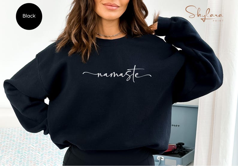 K&ouml;nnte beinhalten: Schwarzer Pullover mit dem Wort "namaste" in wei&szlig;er Kursivschrift.