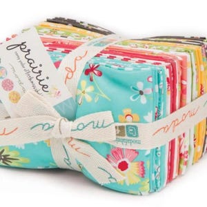 Könnte beinhalten: Ein Bündel bunter Stoffquadrate, gebunden mit einem cremefarbenen Band. Der Stoff zeigt florale Muster in Türkis, Rot und Gelb. Das Band ist mit dem Wort "moda" bedruckt und zu einer Schleife gebunden. Ein Etikett mit dem Wort "prairie" ist angebracht.