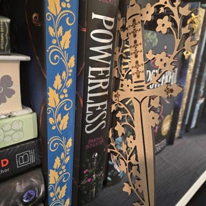 Könnte beinhalten: Ein Bücherstütze aus Holz in Form eines Dolches mit einem Ranken- und Blumendesign. Die Bücherstütze ist braun und hat ein detailliertes Design. Die Bücherstütze steht in einem Bücherregal mit anderen Büchern.