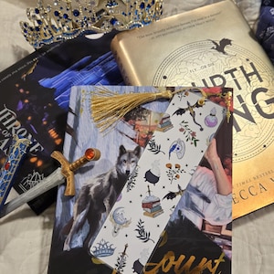 Puede incluir: Una colección de libros de fantasía, una corona decorativa y un marcapáginas. Los libros tienen letras e ilustraciones doradas. El marcapáginas presenta dibujos caprichosos de murciélagos, pociones y libros. La corona es dorada con detalles azules.