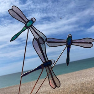 May include: Three stained glass dragonflies with iridescent wings. The dragonflies are mounted on thin metal rods and are arranged in a cluster. The dragonflies are blue, green, and purple.