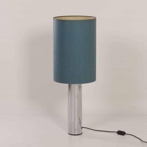 Puede incluir: Una lámpara de mesa de cromo con una pantalla cilíndrica azul. La lámpara tiene un diseño sencillo y una estética moderna.