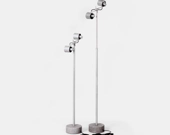 Może przedstawiać: Dwie srebrne lampy podłogowe z regulowanymi reflektorami. Każda lampa ma okrągłą podstawę, wysoki słupek i trzy regulowane reflektory. Lampy są podłączone. Podstawy wykonane są z szarego, teksturowanego materiału. Wysokość jest regulowana.