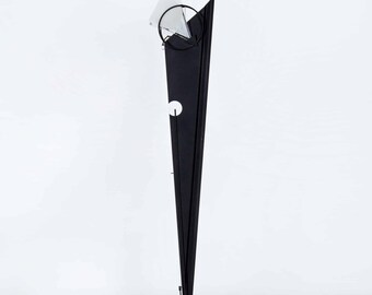 Może przedstawiać: Czarna metalowa lampa stojąca o geometrycznym wzorze. Lampa ma trójkątny klosz i okrągłą podstawę.