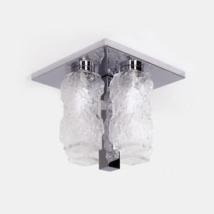 Può includere: Un moderno lampadario da soffitto con due paralumi in vetro smerigliato. I paralumi hanno un aspetto strutturato e organico e sono sospesi da una base quadrata cromata. L'apparecchio è progettato per l'illuminazione dall'alto.