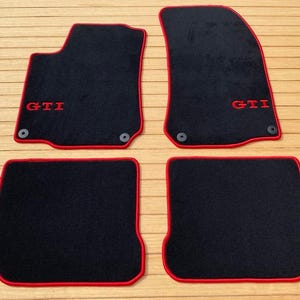Può includere: Set di quattro tappetini per auto neri con bordi rossi. I tappetini presentano la scritta "GTI" ricamata in rosso. Due tappetini sono sagomati per la parte anteriore e due sono rettangolari per la parte posteriore.