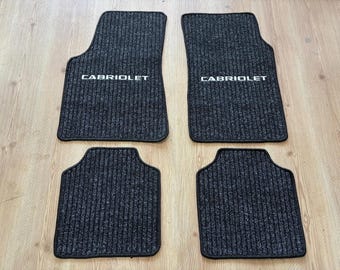 May include: Set of four black car floor mats with a ribbed texture. The word "CABRIOLET" is printed in white on the front mats. The mats are designed to protect the vehicle's interior from dirt and wear.