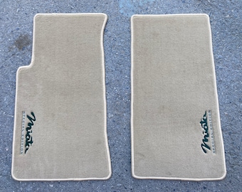 Puede incluir: Dos alfombrillas de coche beige con ribete blanco. Las alfombrillas tienen la palabra "Miata" bordada en verde. La alfombrilla de la izquierda tiene las palabras "Special Edition" bordadas debajo de la palabra "Miata". La alfombrilla de la derecha tiene las palabras "Special Edition" bordadas encima de la palabra "Miata".