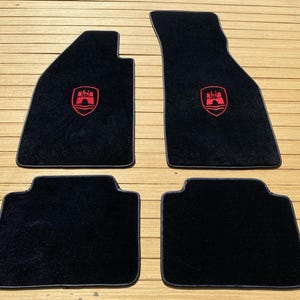 Puede incluir: Un juego de cuatro alfombrillas negras para coche con textura de felpa y ribetes oscuros. Las dos alfombrillas delanteras más grandes presentan un logotipo de escudo bordado en rojo con un diseño de castillo. Alfombrillas para vehículos.
