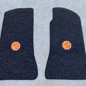 Puede incluir: Dos alfombrillas negras para coche con superficie texturizada. Cada alfombrilla presenta un emblema octogonal rojo y amarillo con las letras "MG". Las alfombrillas tienen un borde negro y están diseñadas para un vehículo.
