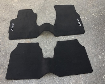 May include: Two black car floor mats. The mats are shaped to fit the footwells of a vehicle. The text "CR-V" is printed in white on the top mat. The mats are made of a soft, carpet-like material.