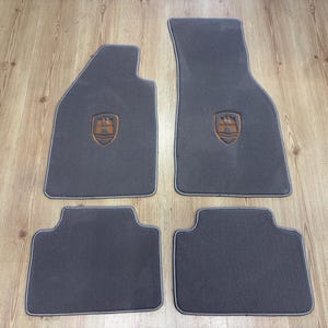 Puede incluir: Un juego de cuatro alfombrillas de coche de color gris oscuro con ribetes de color gris claro en los bordes. Las dos alfombrillas delanteras presentan un logotipo de escudo bordado en naranja y negro, con una torre de castillo sobre líneas.