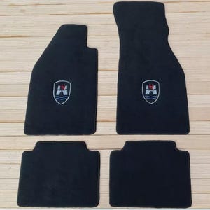 Puede incluir: Juego de cuatro alfombrillas negras para coche. Las alfombrillas tienen forma rectangular y están hechas a medida. Cada alfombrilla presenta un emblema bordado con un diseño de escudo en blanco, rojo y azul.