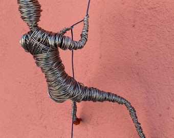 May include: A wire sculpture of a person rappelling down a wall. The sculpture is made of silver wire and is attached to a blue rope. The wall is a light pink color.
