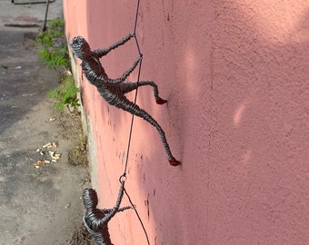 May include: Two wire sculptures of people climbing a pink wall. The sculptures are made of silver wire and are attached to the wall with a blue rope.
