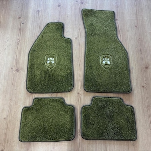 Puede incluir: Un juego de cuatro alfombrillas de coche de color verde oliva con una superficie de moqueta texturizada y ribete oscuro. Dos de las alfombrillas presentan un pequeño emblema en relieve con forma de escudo en el centro, y las otras dos son lisas.