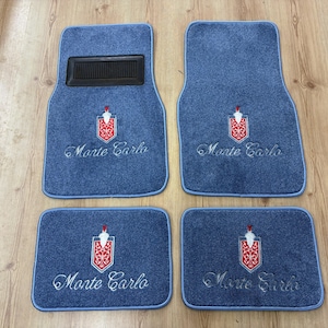 May include: Set of four blue car floor mats. Each mat features the embroidered words "Monte Carlo" in cursive, along with a red and white emblem. The mats have a blue border and a black plastic heel pad.