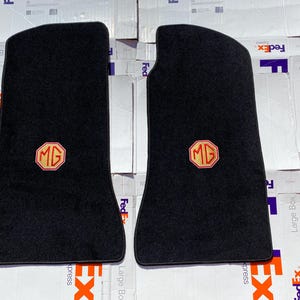 Puede incluir: Dos alfombrillas negras para coche con el logotipo MG dorado y rojo. Las alfombrillas son rectangulares con un borde superior curvado, probablemente para un coche clásico.