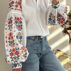 Op de afbeelding: Wit linnen blouse met lange mouwen en een geborduurd bloemenpatroon. De borduursels zijn in tinten rood, oranje, geel, groen, blauw en paars.