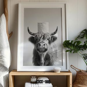 May include: Black and white framed print featuring a Highland cow with a roll of toilet paper on its head. The artwork is displayed on a wooden shelf with a candle, towels, and a decorative brush. A white towel hangs on a wooden hook.