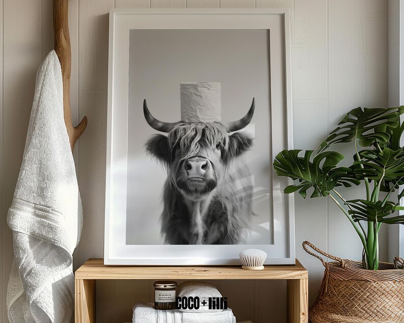 May include: Black and white framed print featuring a Highland cow with a roll of toilet paper on its head. The artwork is displayed on a wooden shelf with a candle, towels, and a decorative brush. A white towel hangs on a wooden hook.