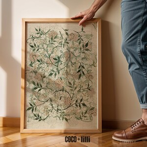 May include: A rectangular framed wall art piece featuring a botanical pattern of green leaves and small pink flowers on a light beige background. The wooden frame is light brown. The text COCO + lilli is printed in black at the bottom center.