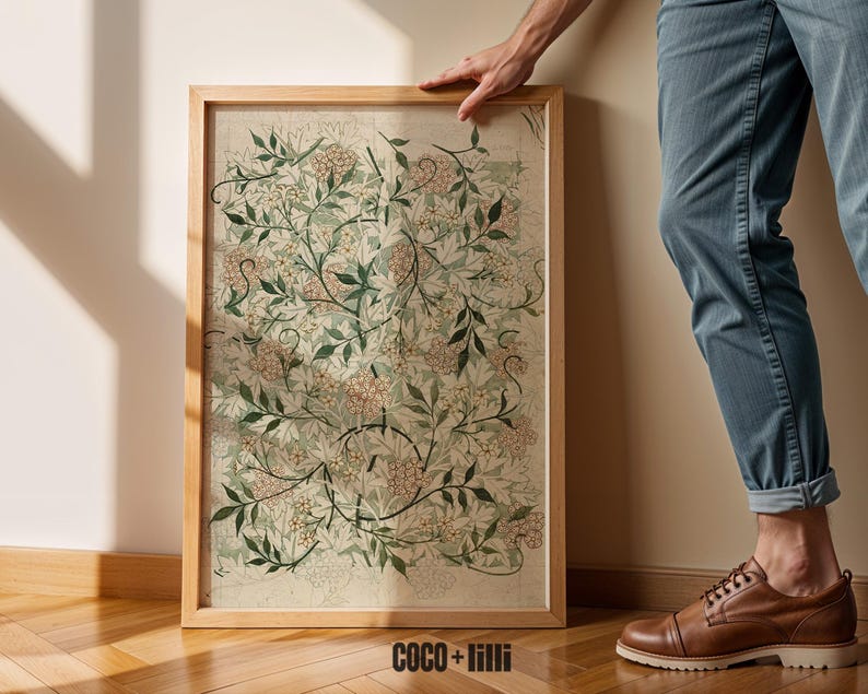 May include: A rectangular framed wall art piece featuring a botanical pattern of green leaves and small pink flowers on a light beige background. The wooden frame is light brown. The text COCO + lilli is printed in black at the bottom center.