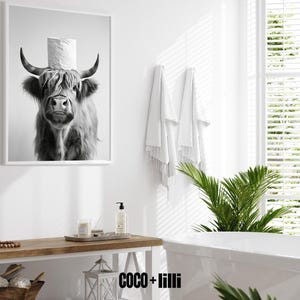 May include: Black and white framed print of a Highland cow wearing a toilet paper roll as a hat. The image is in a bathroom setting with white walls, a wooden table, and a bathtub. The overall theme is humorous bathroom decor.