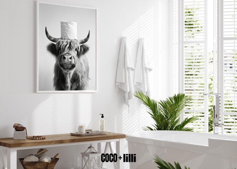 May include: Black and white framed print of a Highland cow wearing a toilet paper roll as a hat. The image is in a bathroom setting with white walls, a wooden table, and a bathtub. The overall theme is humorous bathroom decor.