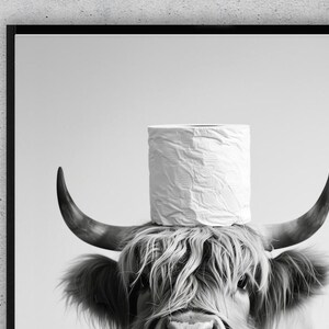 May include: Black and white print of a Highland cow with a roll of white toilet paper balanced on its horns. The cow has long, shaggy fur and curved horns. The image is framed in a black border.