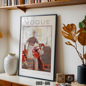 Op de afbeelding: Een ingelijste vintage Vogue tijdschriftomslag met een illustratie van een persoon in een trui met patroon naast een rood-witte auto. Het kunstwerk bevat een kompasroosontwerp en de tekst VOGUE bovenaan de poster.