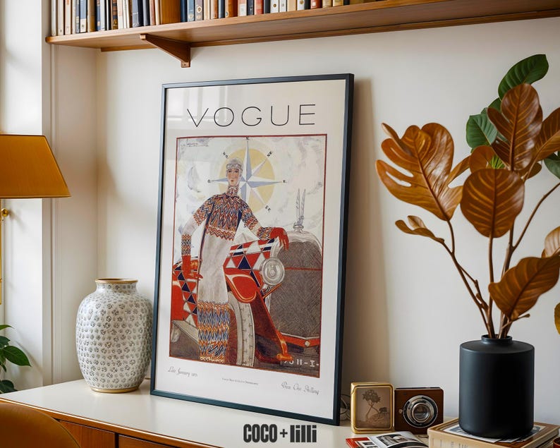 Op de afbeelding: Een ingelijste vintage Vogue tijdschriftomslag met een illustratie van een persoon in een trui met patroon naast een rood-witte auto. Het kunstwerk bevat een kompasroosontwerp en de tekst VOGUE bovenaan de poster.