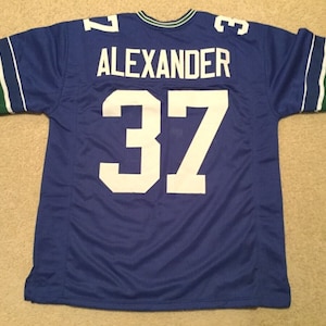 May include: A blue football jersey with white lettering. The name "ALEXANDER" and the number "37" are on the back of the jersey. The jersey has green and white stripes on the sleeves.