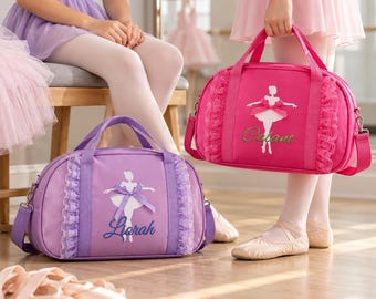 May include: Two duffel bags featuring a ballerina silhouette graphic, lace trim, and a bow. The purple bag is personalized with the name Liorah, and the hot pink bag is personalized with the name Cabant. Both bags have top handles and straps.
