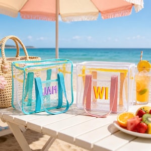 May include: Two clear tote bags with colorful accents and handles sit on a white table. One bag has teal handles and the letters "JAM", the other has pink handles and the letters "WI". A straw bag and a drink are also visible.