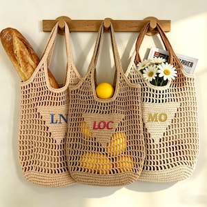 May include: Three beige crocheted tote bags hanging on a wooden rack. One bag holds a loaf of bread, another a lemon and lemons, and the third, flowers and a newspaper. Each bag has a different embroidered initial: LN, LOC, and MO.
