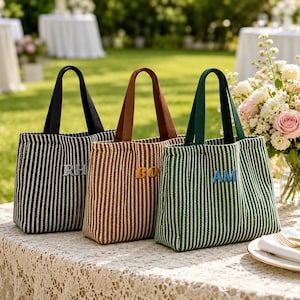 May include: Three striped tote bags in black and white, brown and white, and green and white. Each bag has a different colored handle and embroidered initials: "RH", "BO", and "AM". The bags are sitting on a lace tablecloth.