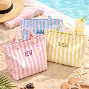 May include: Three striped tote bags in pink, blue, and yellow with white stripes. Each bag has a different embroidered two-letter initial. The bags have clear plastic sides and handles, and are sitting on a sandy beach.