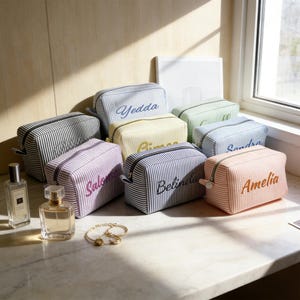 Puede incluir: Una colección de neceseres personalizados en varios colores, como rosa, lavanda y azul, con nombres bordados. Las bolsas tienen un patrón de rayas y cierre de cremallera. También se ven frascos de perfume y una pulsera.
