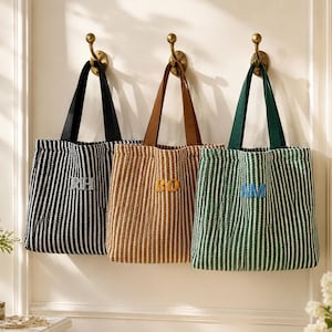 May include: Three rectangular tote bags with vertical stripes hanging on brass hooks. The left bag is black and white with the letters RH, the center bag is tan and white with BO, and the right bag is green and white with the letters AM embroidered.