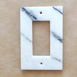 May include: A rectangular, white marble light switch cover with black veining. The cover has a single rectangular opening in the center and two small holes for mounting. The background is a light brown surface.