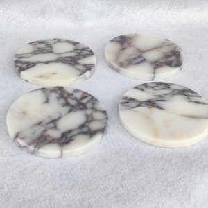 Pu&ograve; includere: Quattro sottobicchieri rotondi in marmo con sfondo bianco e venature grigie e viola.