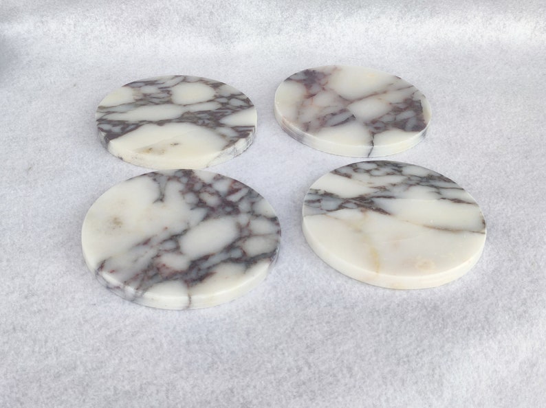 Pu&ograve; includere: Quattro sottobicchieri rotondi in marmo con sfondo bianco e venature grigie e viola.