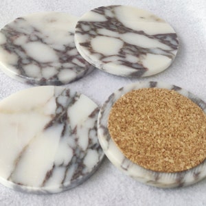 Pu&ograve; includere: Set di quattro sottobicchieri rotondi in marmo con un motivo a marmo bianco e grigio. Un sottobicchiere ha un retro in sughero.